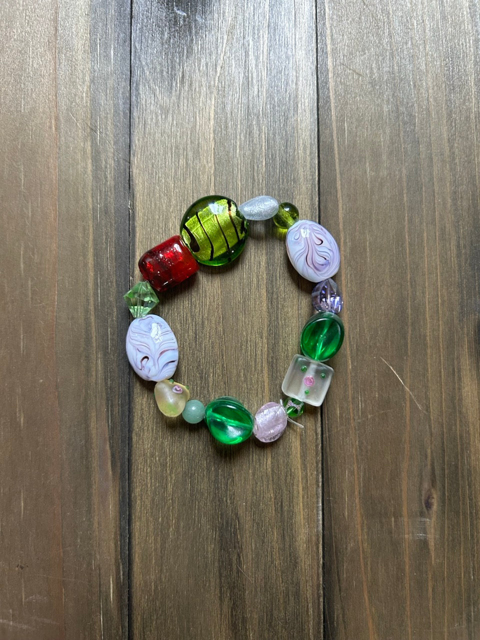 A handmade beaded stretch bracelet featuring a variety of colorful antique glass beads. Includes a variety of shapes and sizes of green, white, red, and clear beads, displayed on a dark wood background.