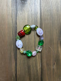 A handmade beaded stretch bracelet featuring a variety of colorful antique glass beads. Includes a variety of shapes and sizes of green, white, red, and clear beads, displayed on a dark wood background.