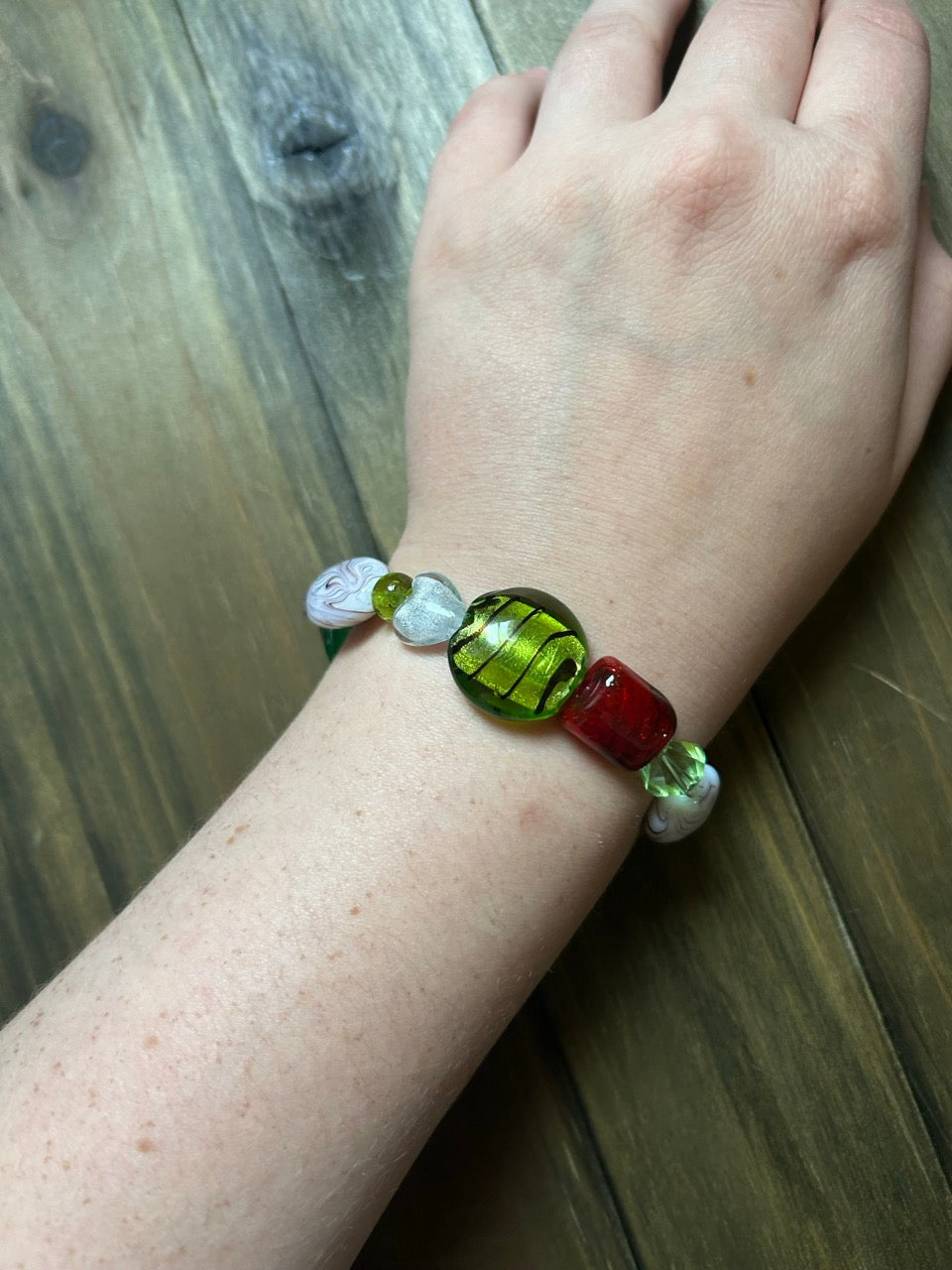 A close-up of a person's wrist wearing a handmade beaded bracelet with a large green glass bead, a red glass bead, and smaller white and green beads, set against a dark wood background.