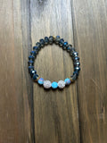 A beaded stretch bracelet featuring faceted dark blue-gray beads, two light blue round beads, and two silver-toned crystal-encrusted round beads, displayed on a dark wood background.