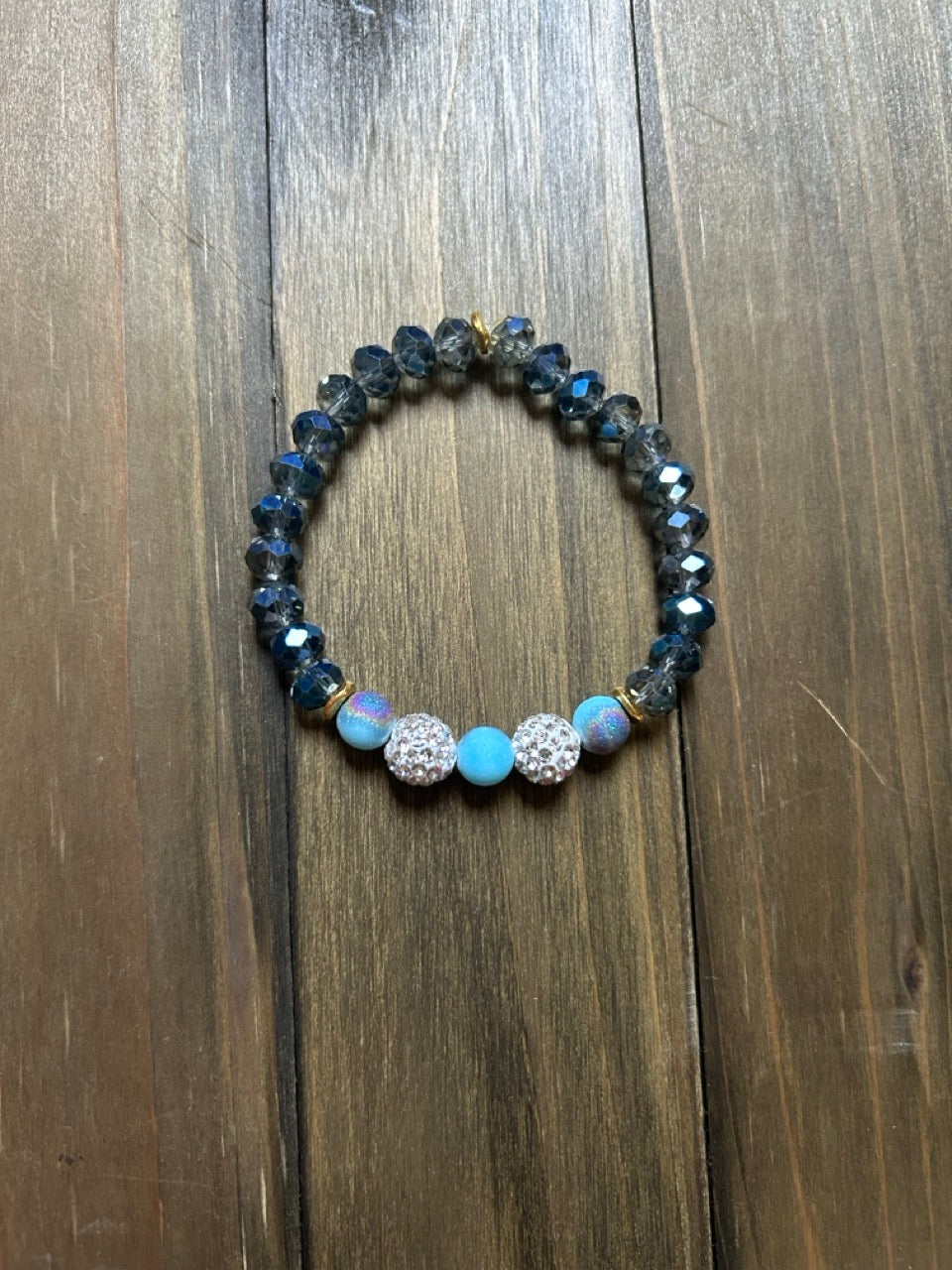 A beaded stretch bracelet featuring faceted dark blue-gray beads, two light blue round beads, and two silver-toned crystal-encrusted round beads, displayed on a dark wood background.