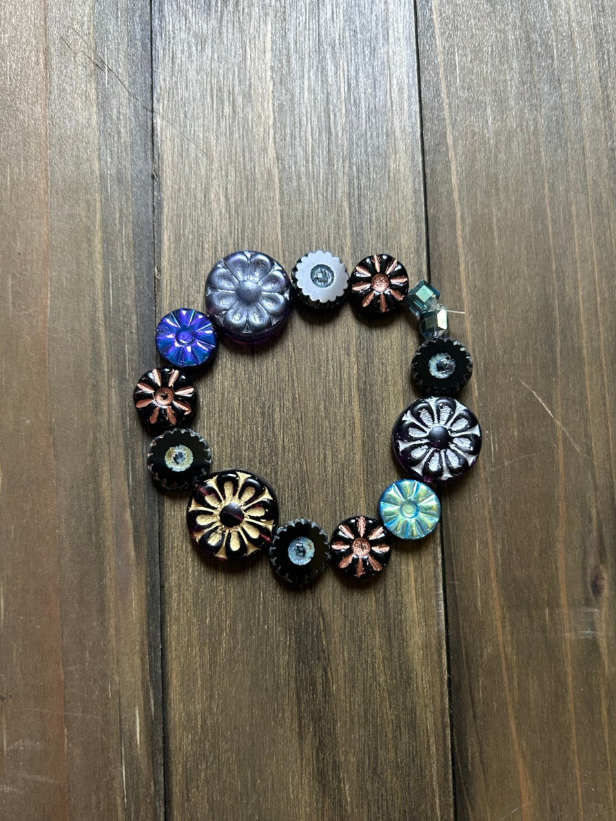 A handmade stretch bracelet made of colorful black, blue, gold, and silver  flower-shaped beads, displayed on a dark brown wooden surface.