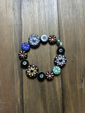 A handmade stretch bracelet made of colorful black, blue, gold, and silver  flower-shaped beads, displayed on a dark brown wooden surface.