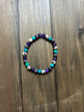 A beaded stretch bracelet featuring purple, turquoise, gray, and white-and-black spotted beads, displayed on a dark brown wooden surface.