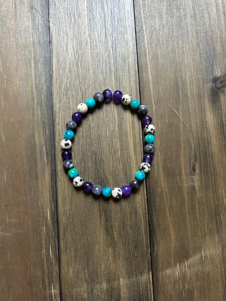 A beaded stretch bracelet featuring purple, turquoise, gray, and white-and-black spotted beads, displayed on a dark brown wooden surface.