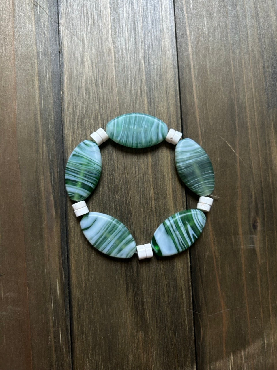 Green and white striped glass beaded bracelet with white spacer beads made of white shell. On a dark wood plank background.