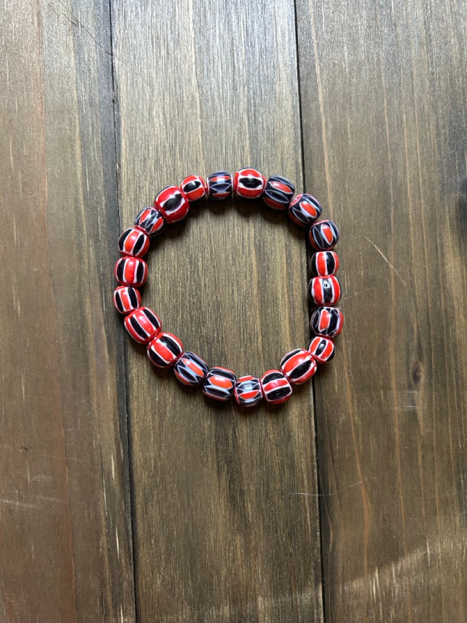 A beaded stretch bracelet made of glossy, round beads with a red, black, and white striped pattern. Displayed on a dark brown wooden surface.