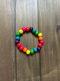 A wooden beaded stretch bracelet arranged in a rainbow sequence of red, orange, yellow, green, blue, and purple beads, displayed on a dark brown wooden surface.