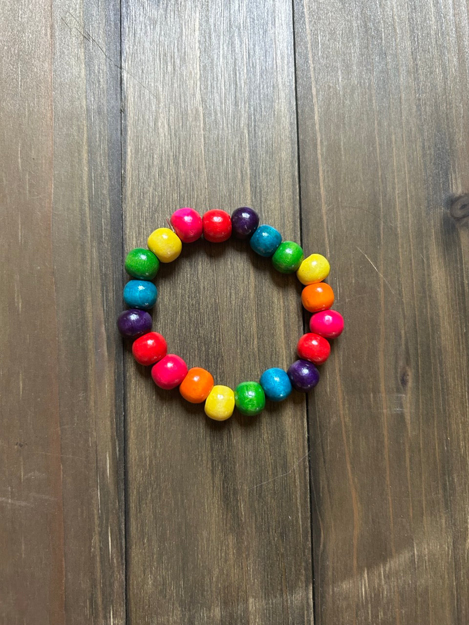 A wooden beaded stretch bracelet arranged in a rainbow sequence of red, orange, yellow, green, blue, and purple beads, displayed on a dark brown wooden surface.