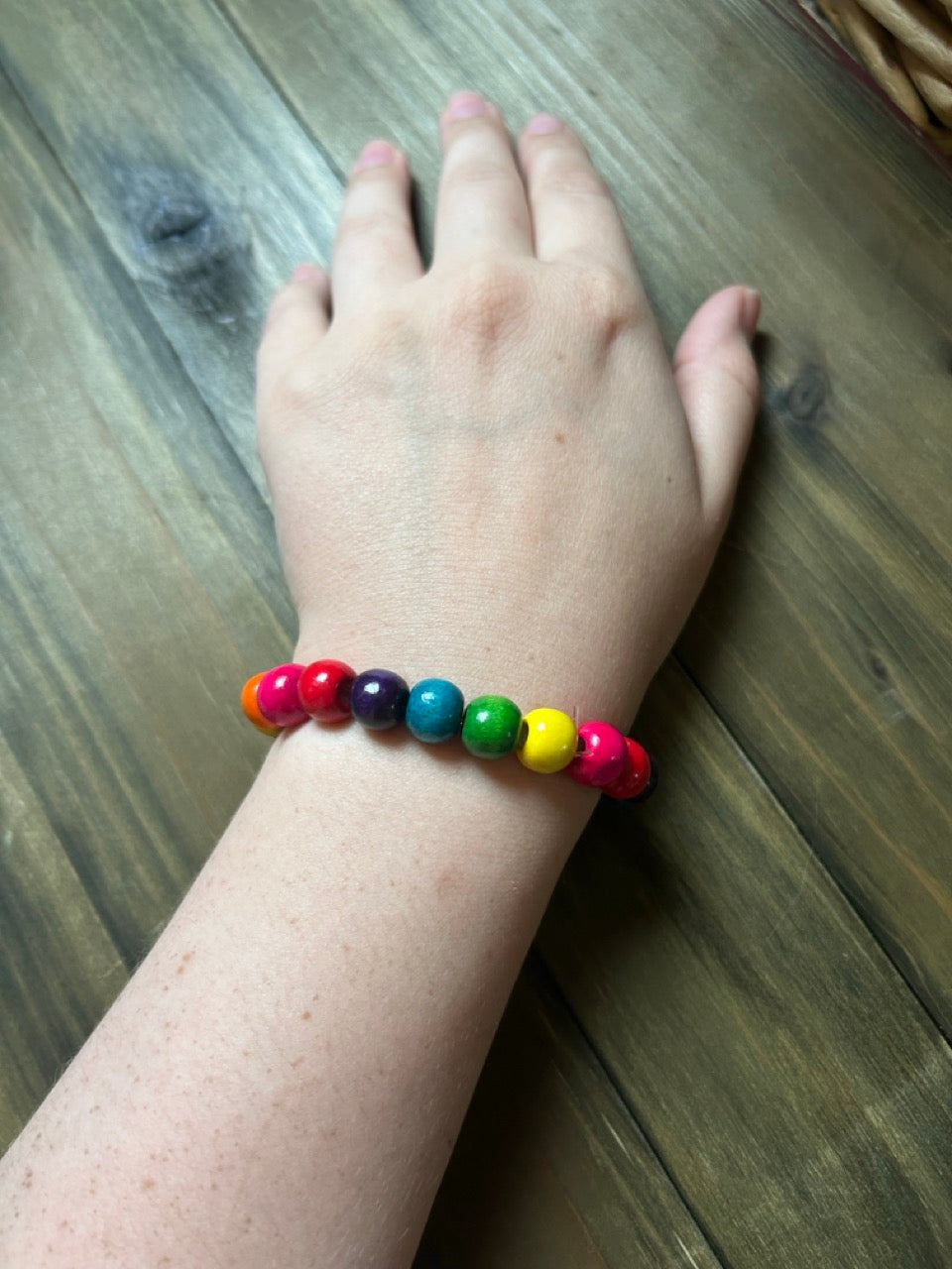 A multicolor beaded stretch bracelet worn on a person's wrist, featuring rainbow-colored wooden beads, shown against a dark wood plank background.