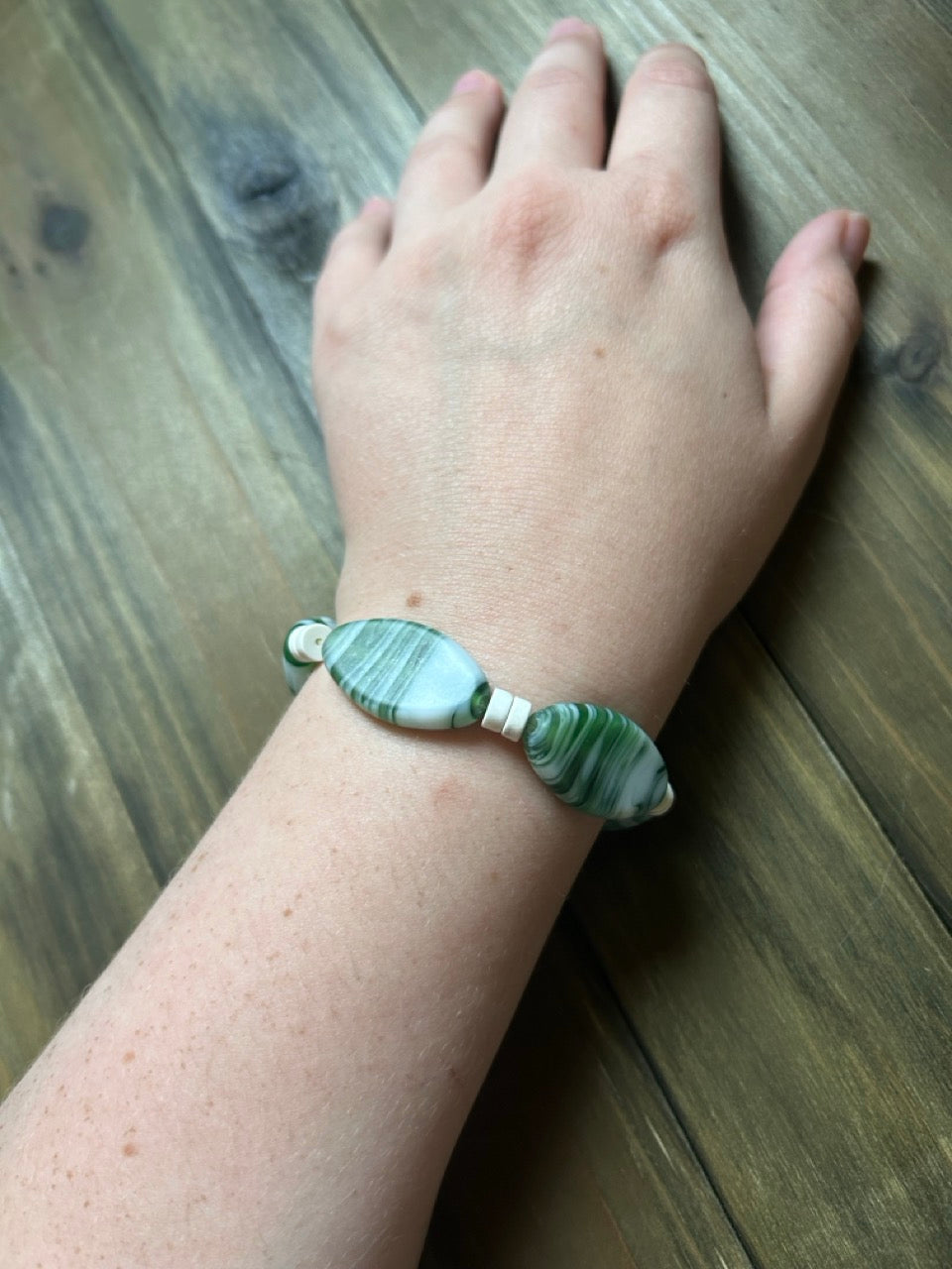 A person's wrist wearing a beaded bracelet made of green and white striped, football-shaped glass beads. There are small white spacer beads in between the glass beads. The hand is resting on a dark wood plank surface.