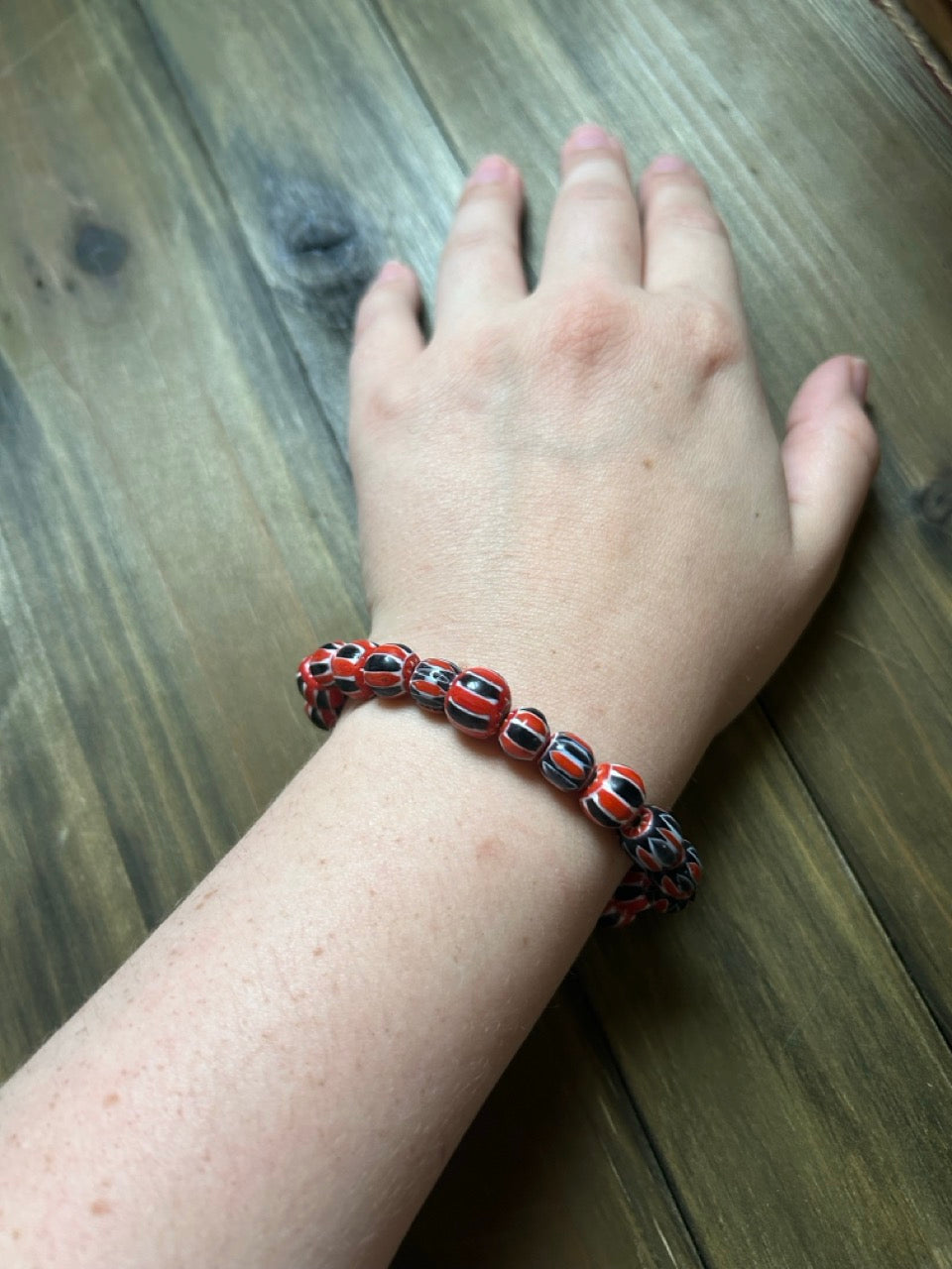 A close-up of a person's hand and wrist wearing a beaded stretch bracelet with red, black, and white patterned beads, resting on a dark wood surface.