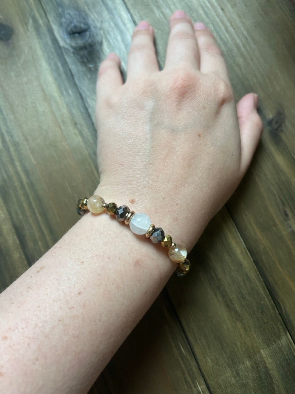 A beaded stretch bracelet with natural stone beads and metallic spacer beads worn on a person's wrist, shown against a dark wood plank background.