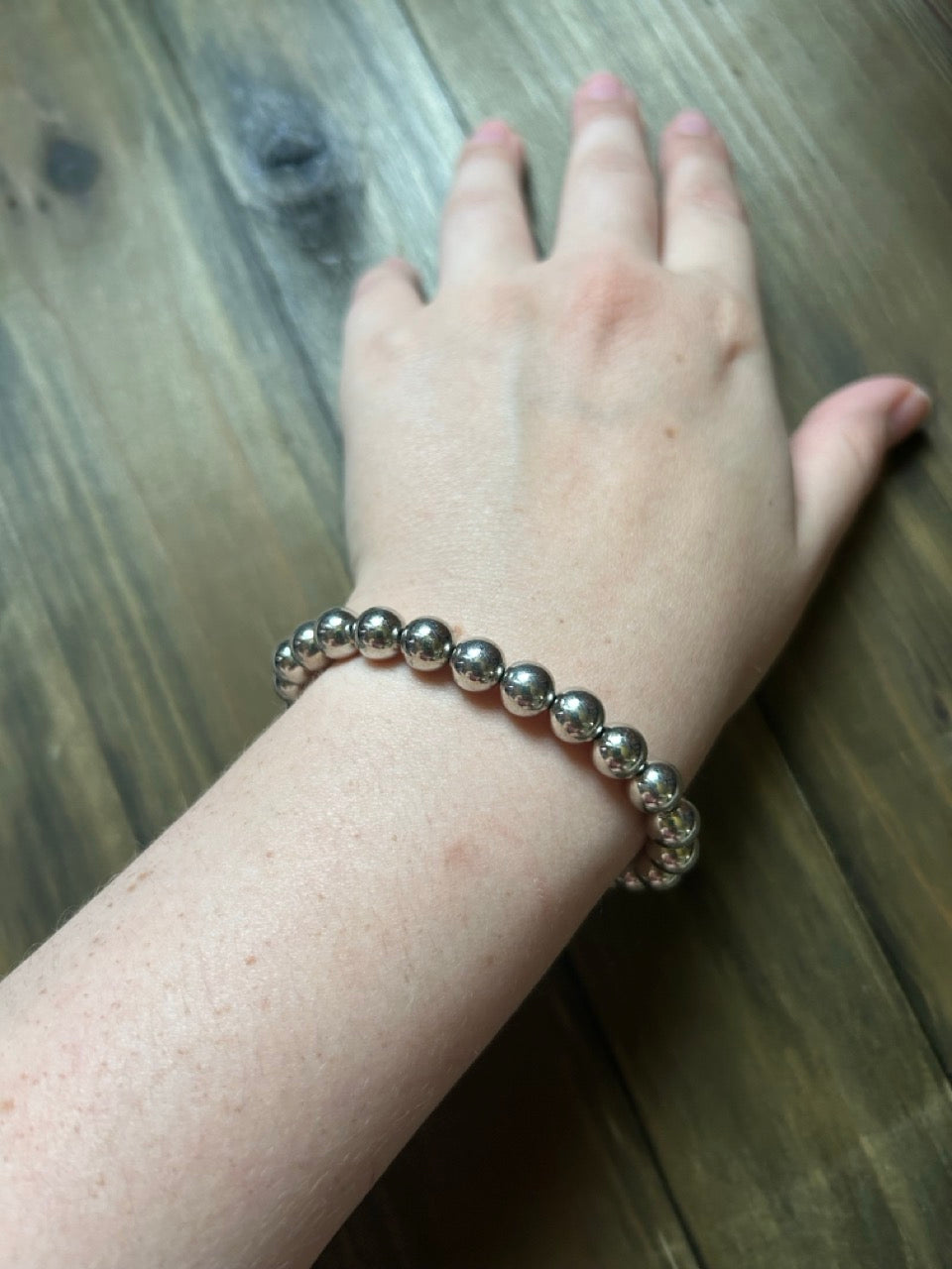 A close-up of a person's wrist wearing a silver beaded bracelet, resting on a dark wood surface.