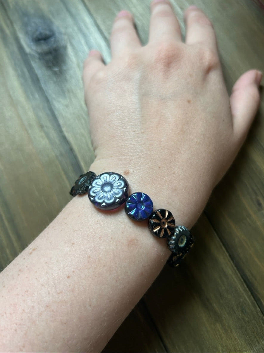 A hand resting on a dark wood plank surface wearing a black and blue Czech glass flower bracelet.