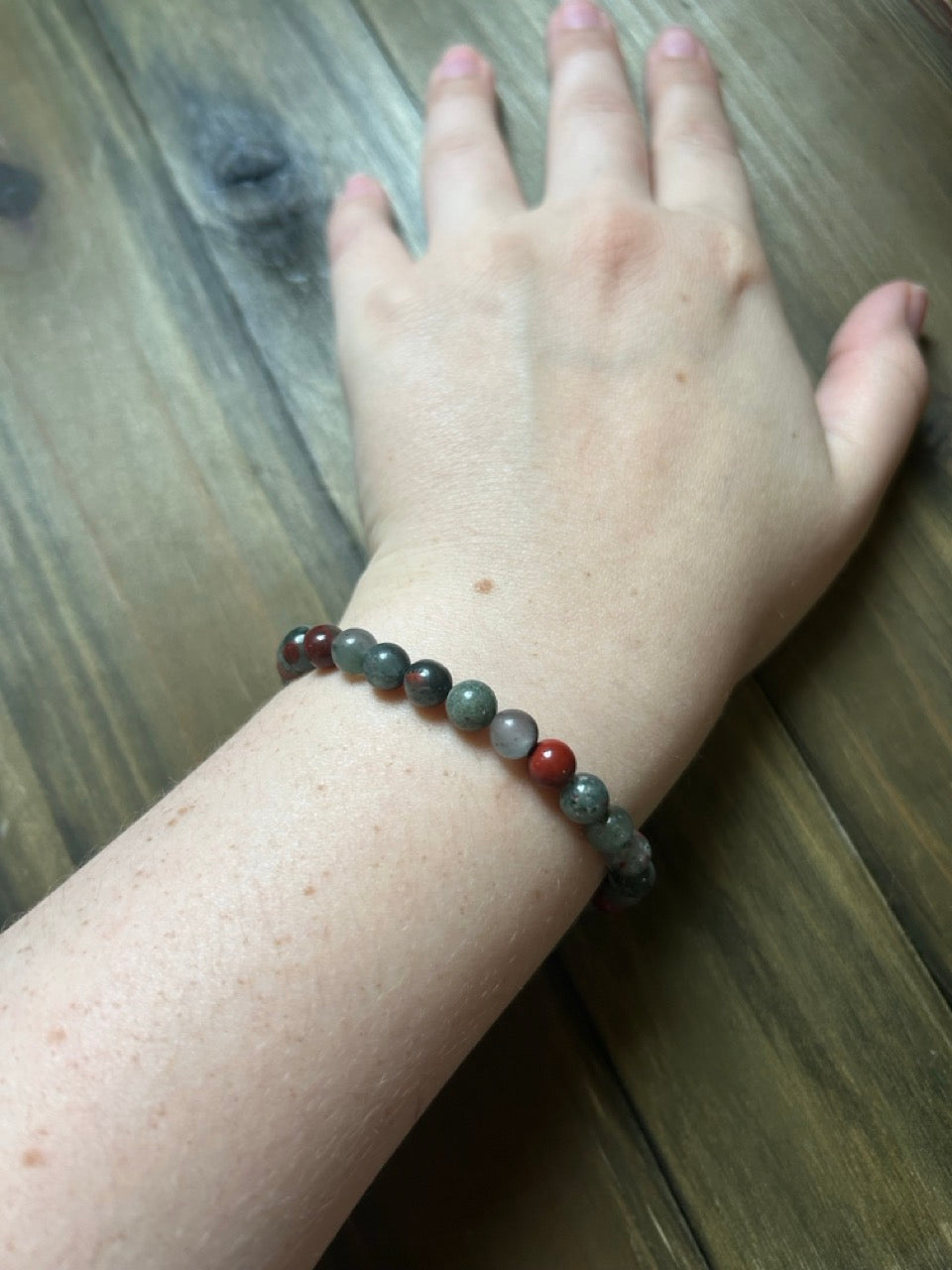 A beaded bracelet worn on a person's wrist, featuring a mix of gray and red marbled, polished beads. The hand is resting against a dark wood plank background.