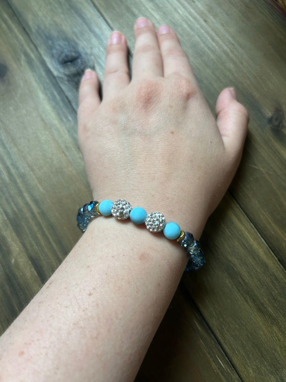 A close-up of a person's wrist wearing a beaded stretch bracelet with light blue beads, silver filigree, and dark gray-blue beads, resting on a wooden surface.