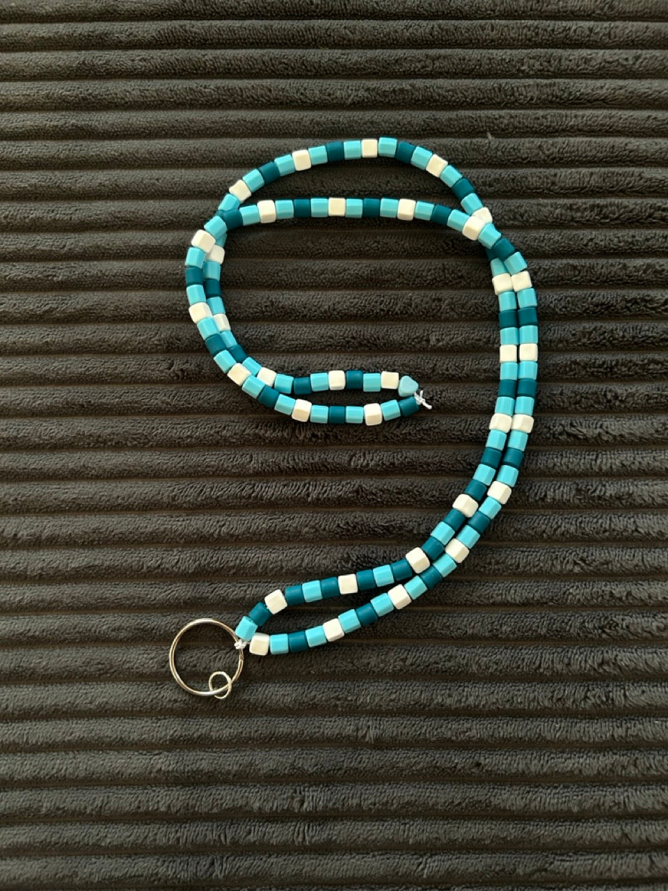 A beaded necklace with teal, light blue, and white square polymer clay beads, featuring a silver-toned split ring clasp, displayed on a dark textured background. 