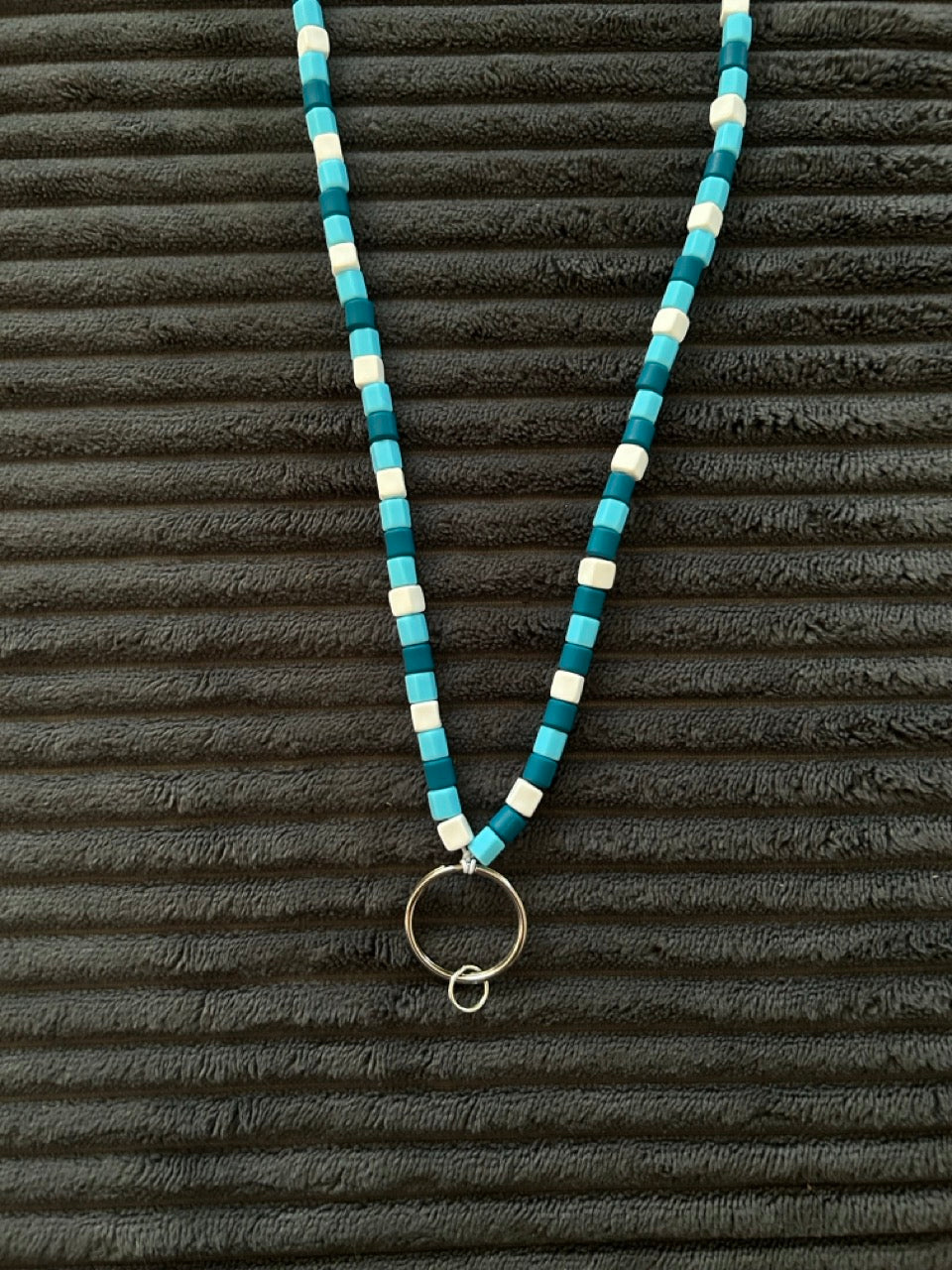 A beaded necklace with light blue, teal, and white cylindrical beads, featuring a silver-toned circular ring pendant, displayed on a dark ribbed background.