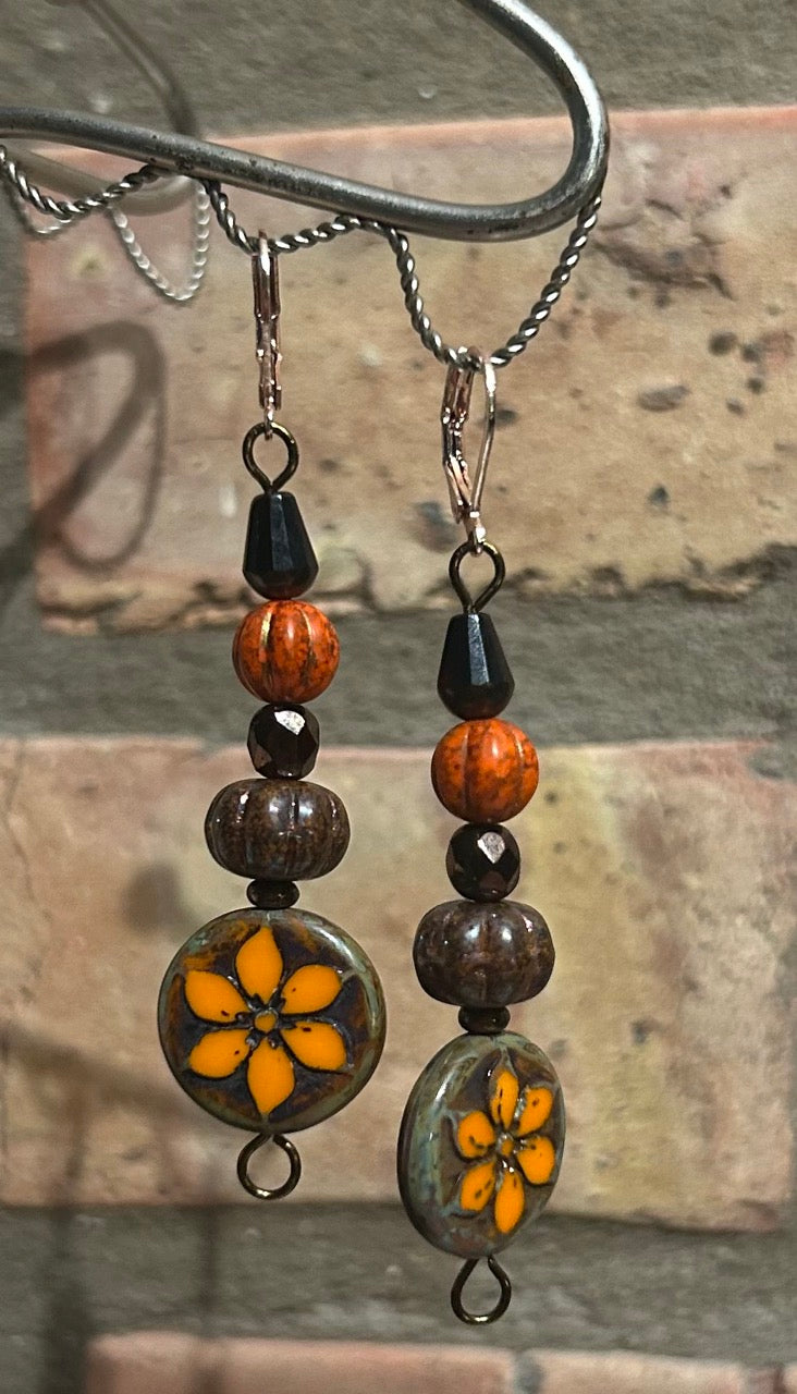 Brown, dark brown, and orange beaded dangle Czech glass earrings. The bottom of the dangle has a bright orange flower in the center. Earring are hanging from a silver hook in front of a light brown brick background.