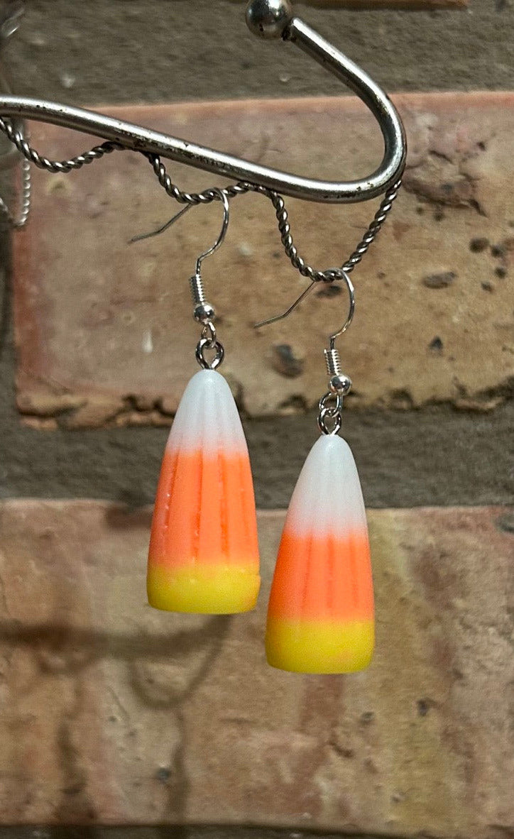 Life size candy corn earrings hanging from a silver hook in front of a light brown brick background.