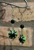 Silly black spiders on green glass beads, dangling with clear and black beads. Earring are hanging from a silver hook in front of a light brown brick background.